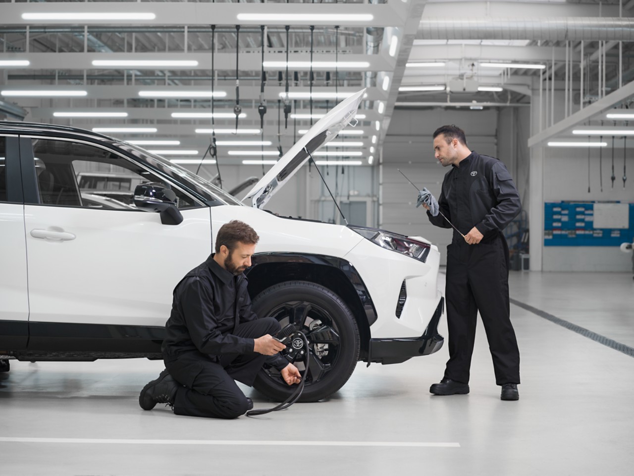 Deux techniciens réparent un Toyota RAV4 blanc, dont le capot est relevé. L’un s’agenouille pour inspecter la pression des pneus, l’autre le niveau d’huile.